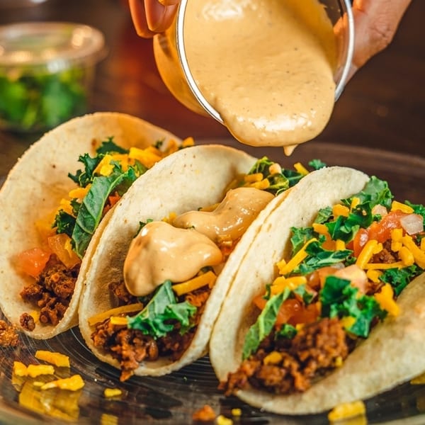 Three tacos filled with meat, cheese, and greens, being drizzled with sauce on a clear plate.