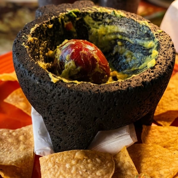 Fresh guacamole in a molcajete with an avocado pit, surrounded by tortilla chips on a red plate.