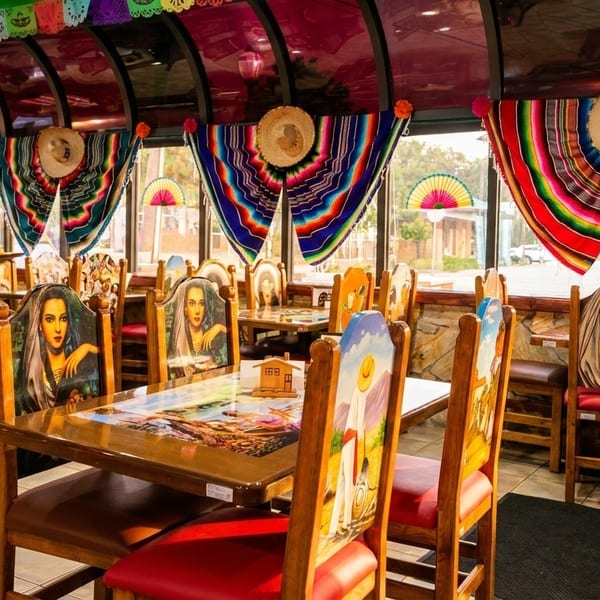 Vibrantly decorated Mexican restaurant dining room with painted chairs, colorful serapes, and a server walking through.