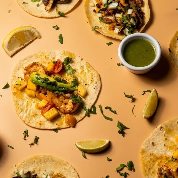 Assortment of tacos with various fillings, salsas, and lime wedges on a light orange surface.