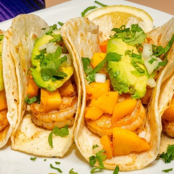 Four shrimp tacos with mango, avocado, and cilantro on a white plate with a lemon wedge.
